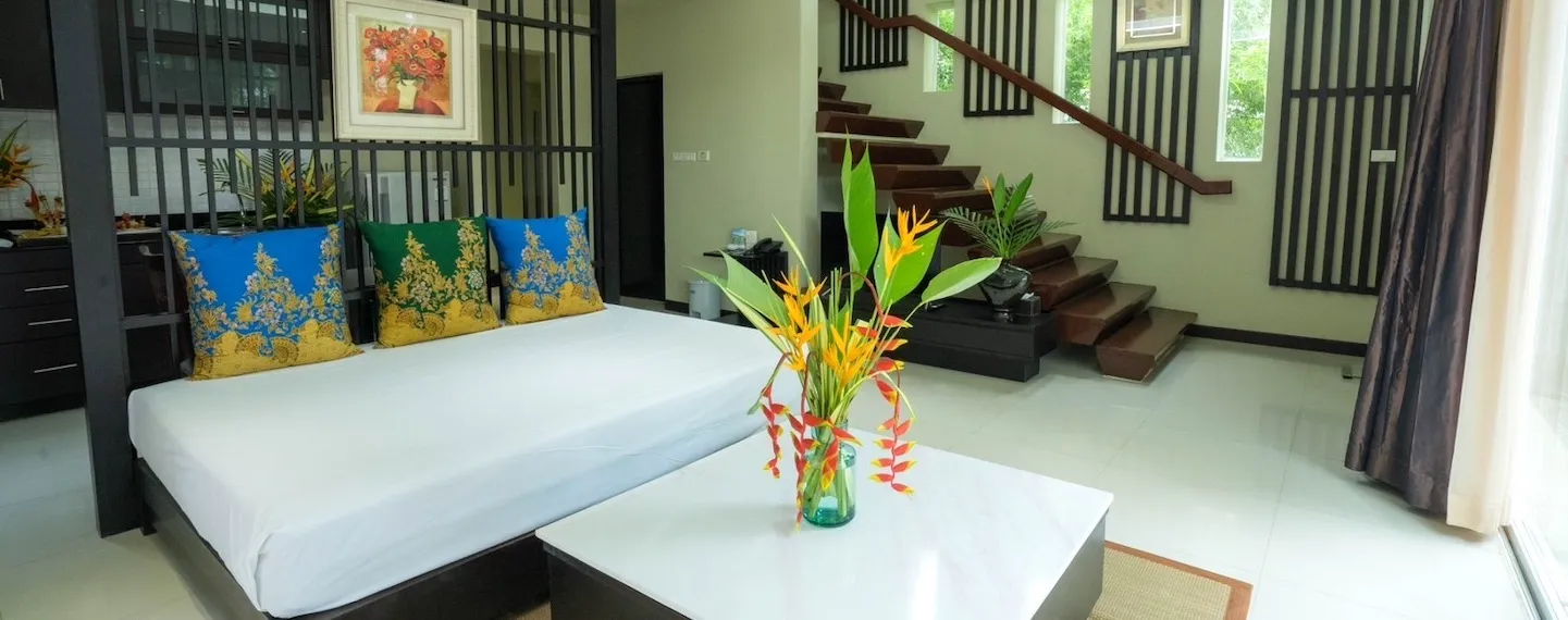 Stylish villa interior with a large bed, cushions, glass coffee table with tropical flowers, and a wooden staircase leading to the upper level
